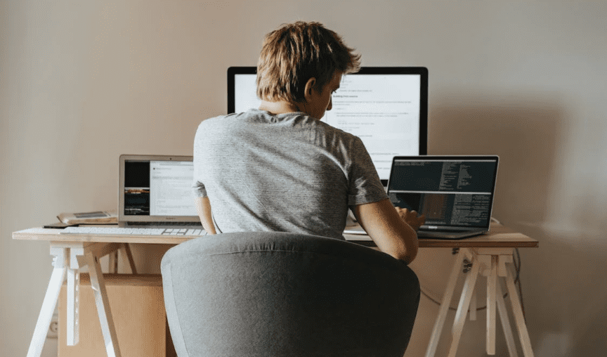 Guy seated at his working desk at home with 2 laptops and 1 monitor