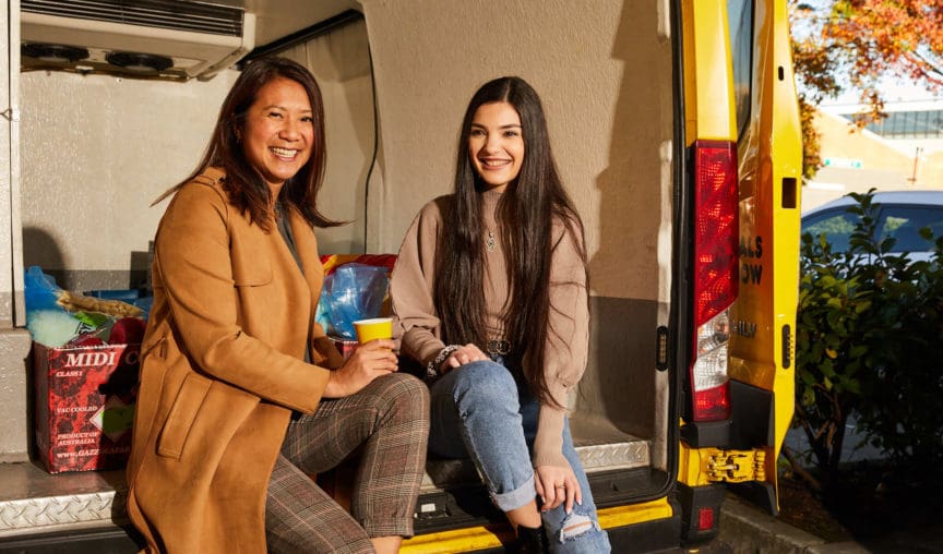 Two women sitting on the back of a van drinking coffee