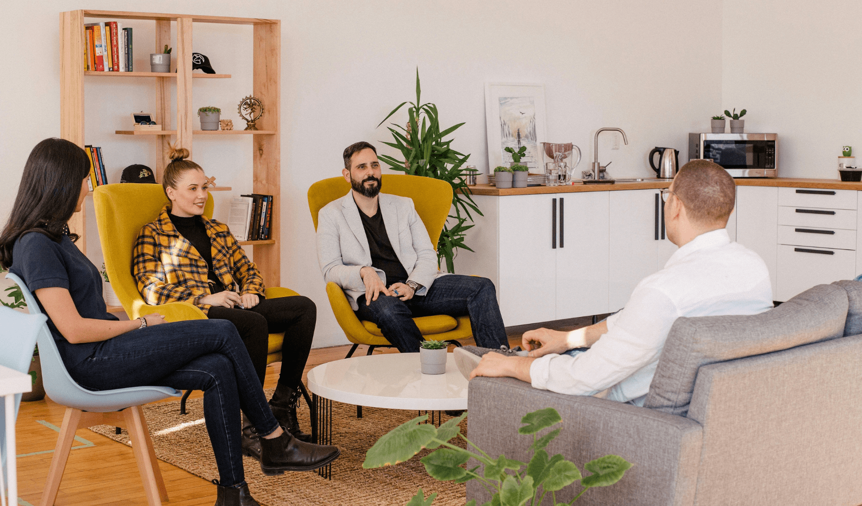 A group of people sit in a casual meeting room, on comfy chairs around a coffee table